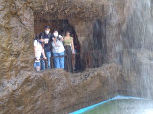 Kilka mężczyzn i kobiet w maseczkach ochronnych. Przebywają w kamiennej grocie. Przed nimi wodospad - ściana wody oraz zbiornik wodny.